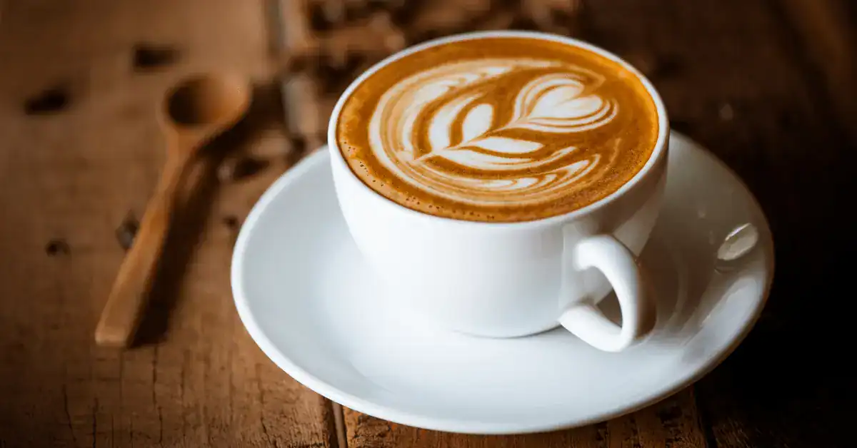 A latte with leaf latte art in a white cup and saucer on a wooden table