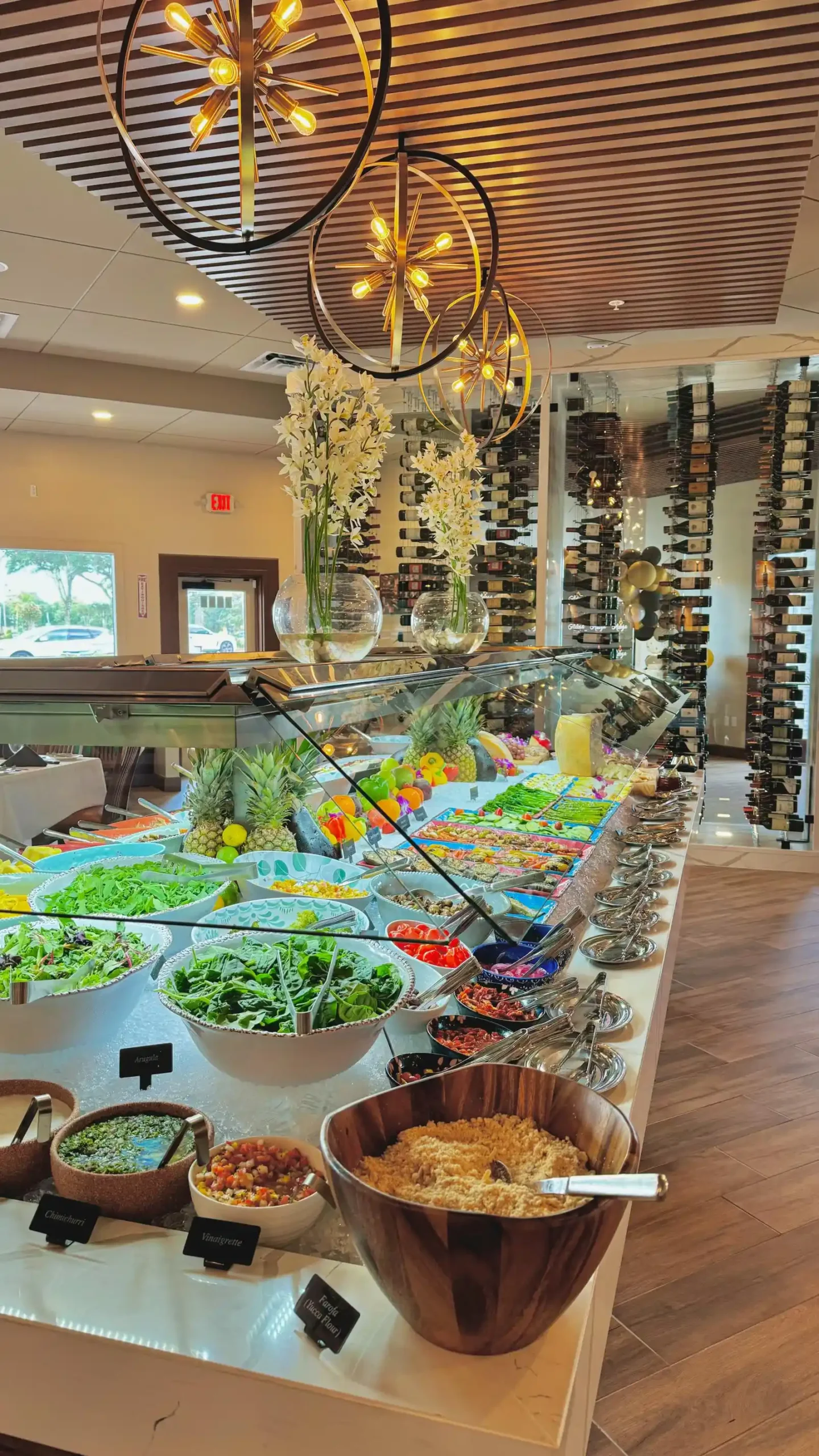 The Gourmet Table at Adega Gaucha Kissimmee with salads and sides beneath gold chandeliers and the wine wall